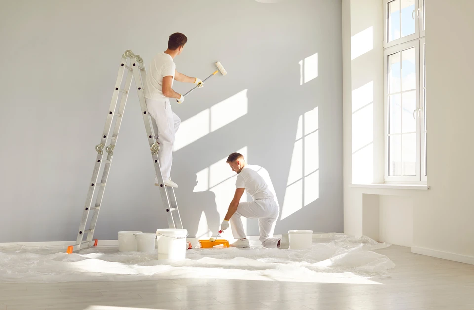 Équipe de peintres professionnels en vêtements blancs peignant un mur gris dans une pièce vide avec rouleau de peinture