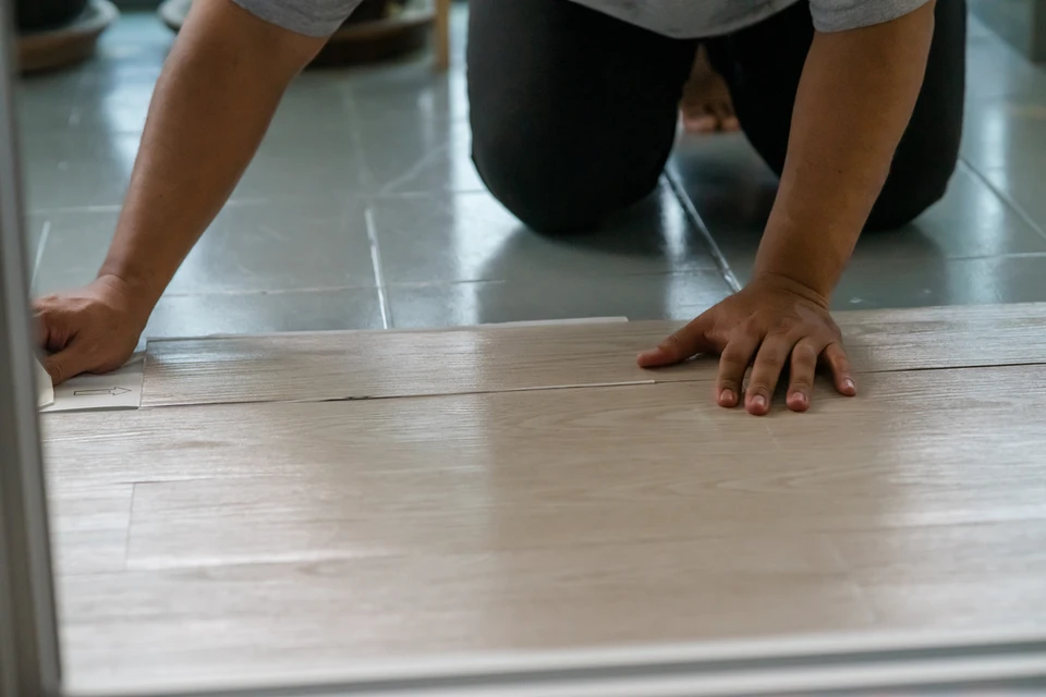 Une personne en vêtements de travail installe du parquet flottant dans un intérieur résidentiel, agenouillée sur carrelage existant